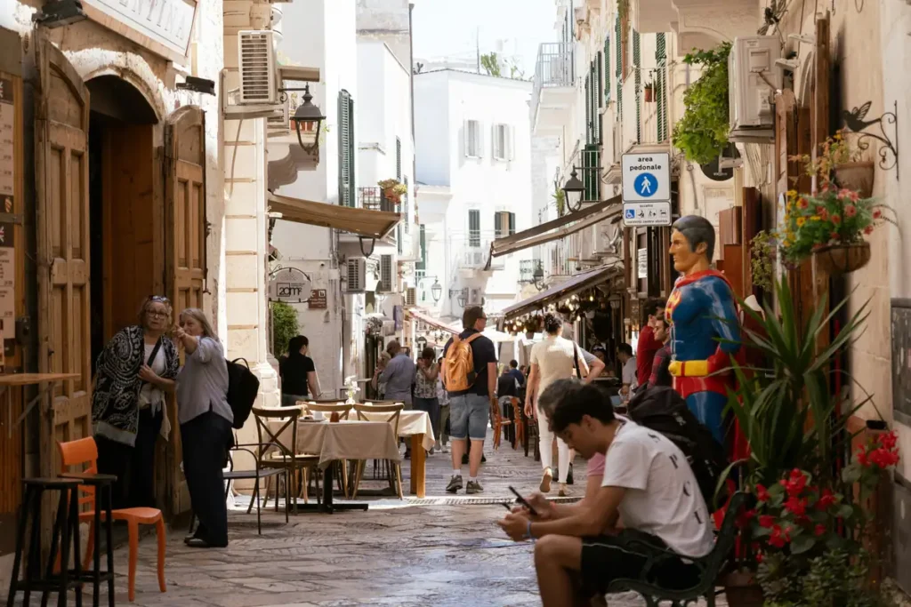 A busy pedestrian alley in Bari's old town with whitewashed buildings, outdoor café tables, tourists walking, flowering plants, and a Superman mannequin outside a shop.

