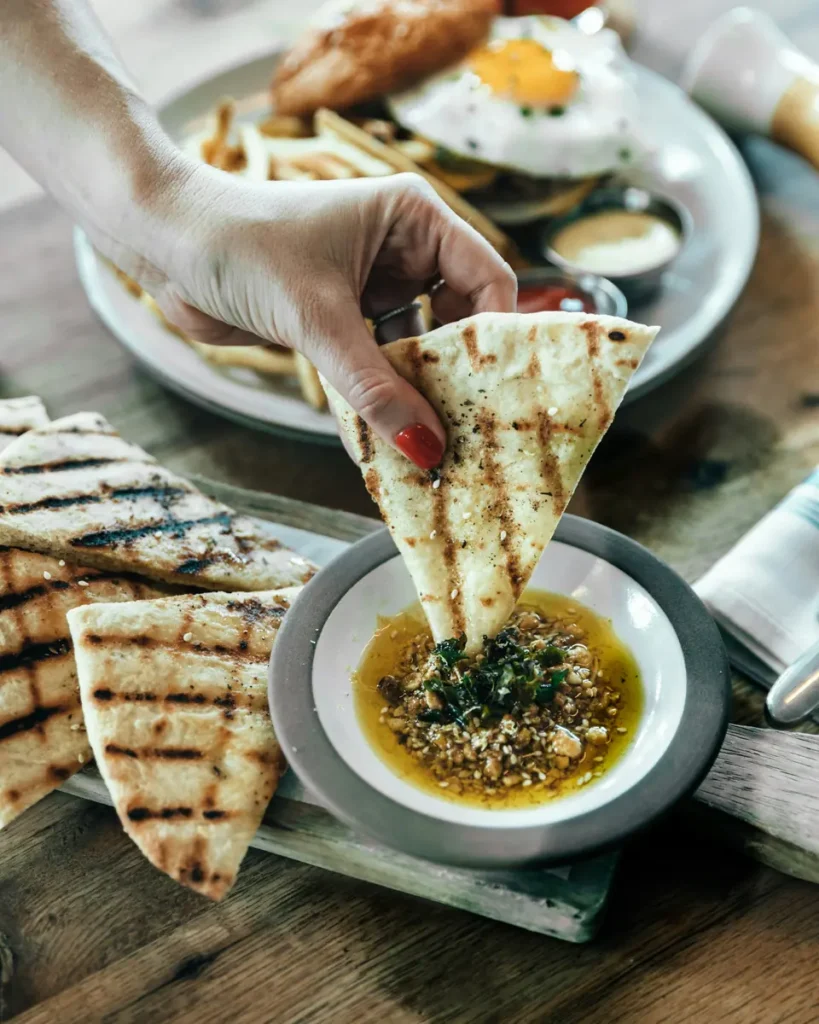 A hand dipping a grilled focaccia slice into a small bowl of herb and olive oil sauce on a wooden board, with a full breakfast plate visible in the background.
