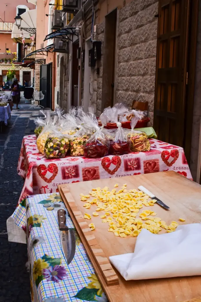 Freshly made orecchiette pasta scattered on a wooden cutting board on a street stall in Bari's old town, with colorful packaged pasta varieties displayed on a table in the background.You said: ye image mai ny article k andr lgani hai mjy is k sth caption r alt text likh k dy do.