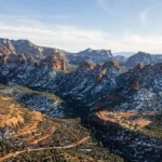 Aerial view of Sedona in winter with snow-dusted red rock formations and green pine trees under a clear blue sky